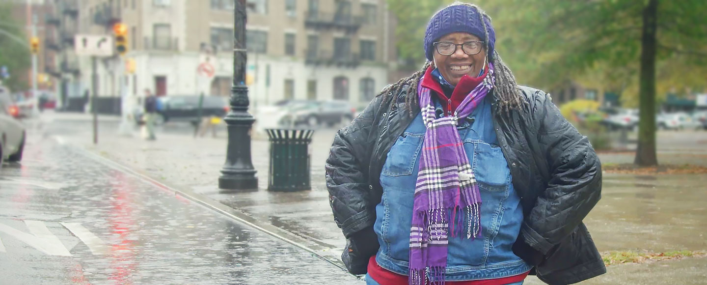 A woman with long braided hair, in a winter coat, hat, and scarf, stands smiling confidently in a New York City streetscape.