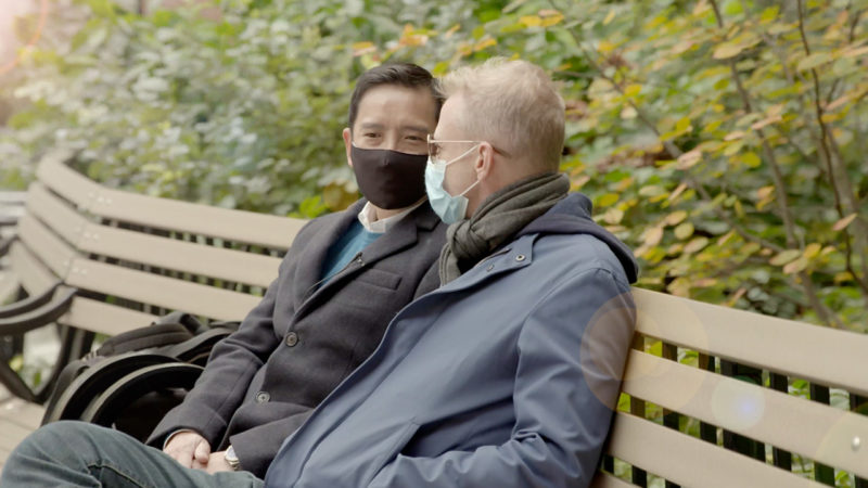 Two men sit on bench talking, one is facing the camera