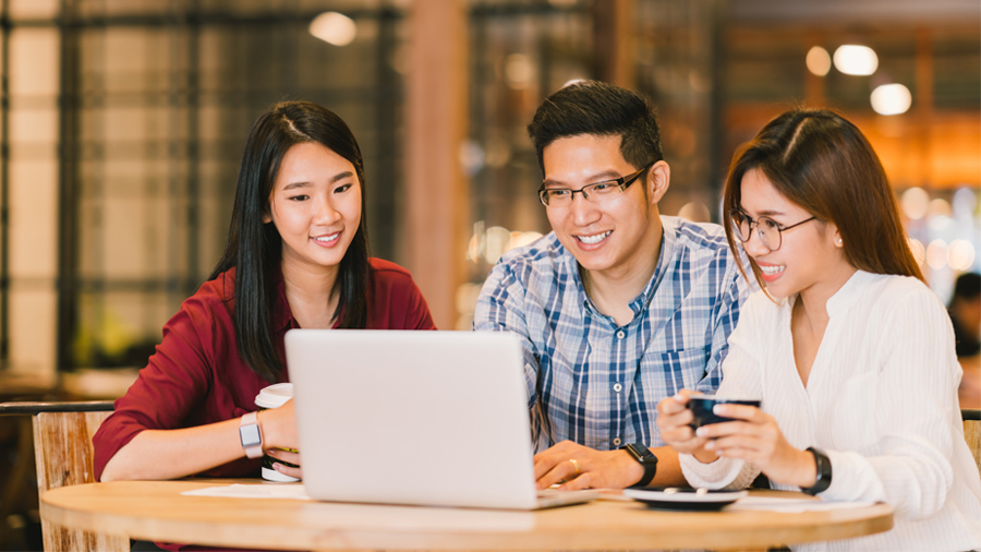 Three young adults are sitting around a laptop and smiling.