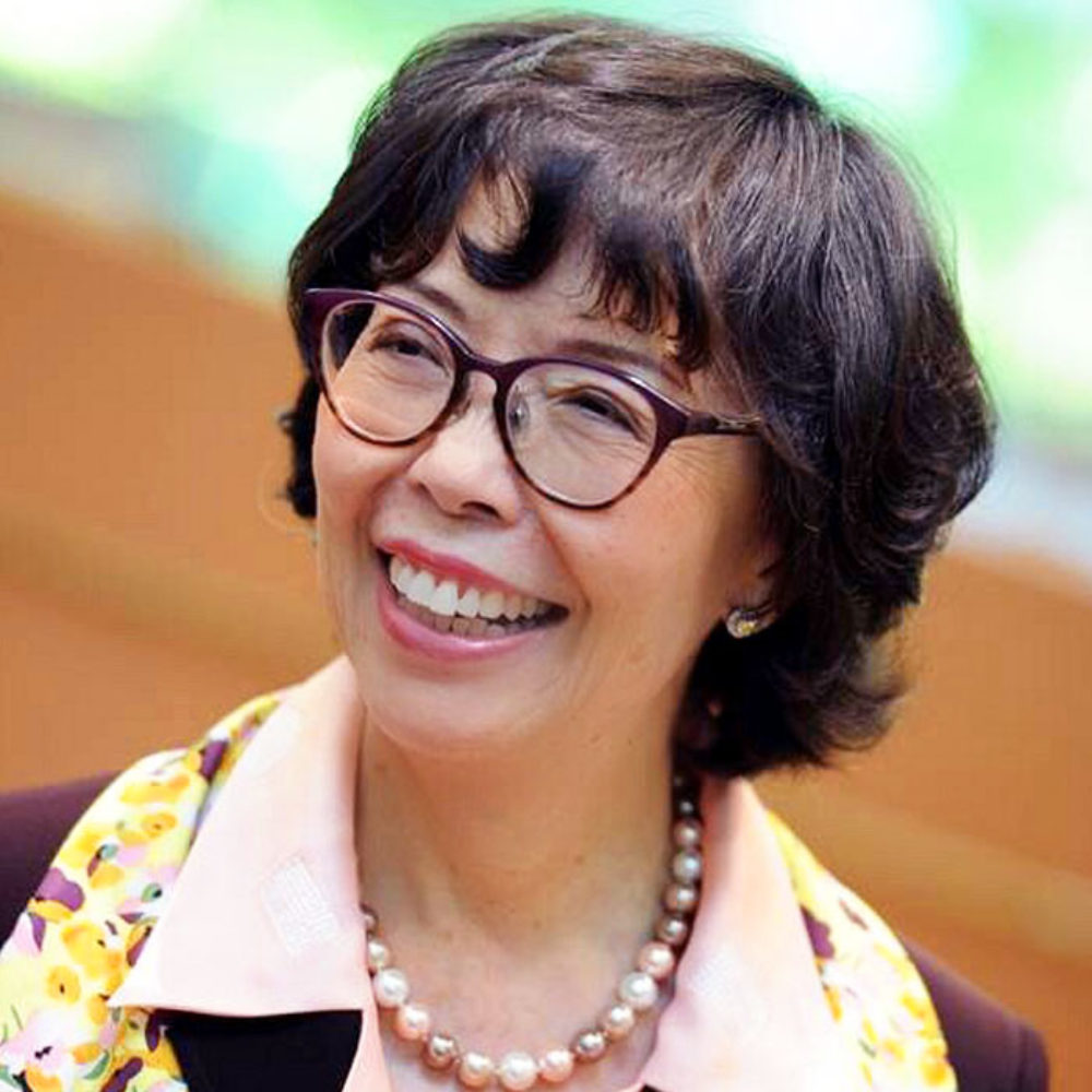 A smiling woman with glasses wearing a pink collared shirt and pearl necklace.