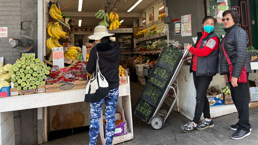 Three people are standing outside of a small market with a fruit stand.