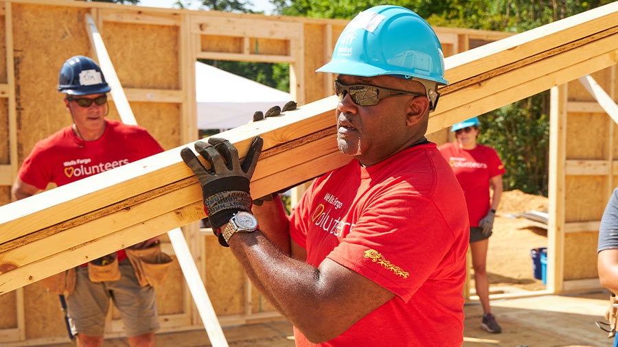 A man in a hardhat carries wood planks at a construction site.