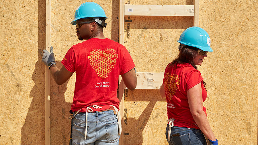 Two people in hardhats stand in front of the wall of a partially built home.