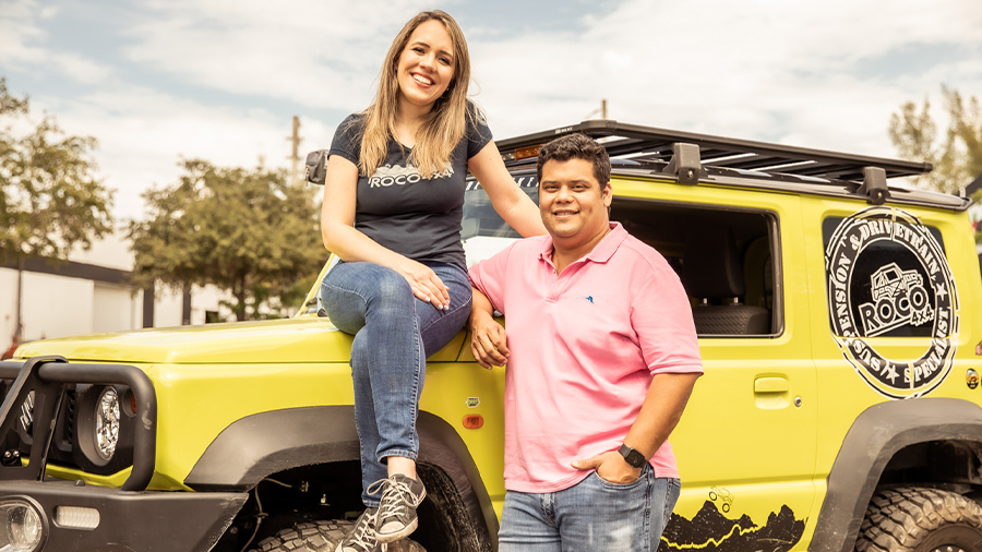 A woman sits on the hood of a yellow SUV while a man stands beside her leaning against the vehicle. Both are smiling for the camera.