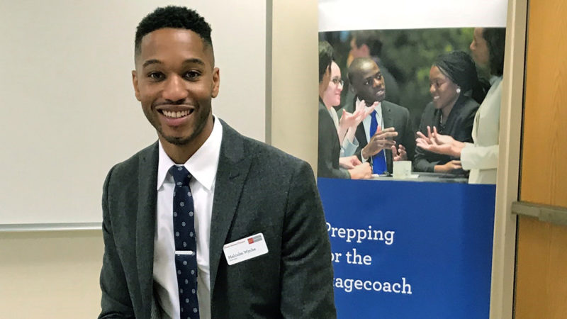 Malcolm Wyche, smiling and wearing a suit, stands in front a banner that reads 