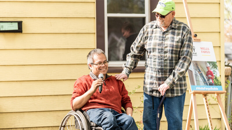 A man in a wheelchair sits in front of a house and holds a microphone. A man with a cane stands next to him smiling.