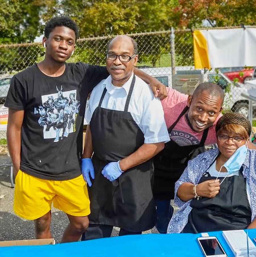 A man wearing an apron has a younger person standing next to him with his arm around him, and two other smiling people on the other side of him.