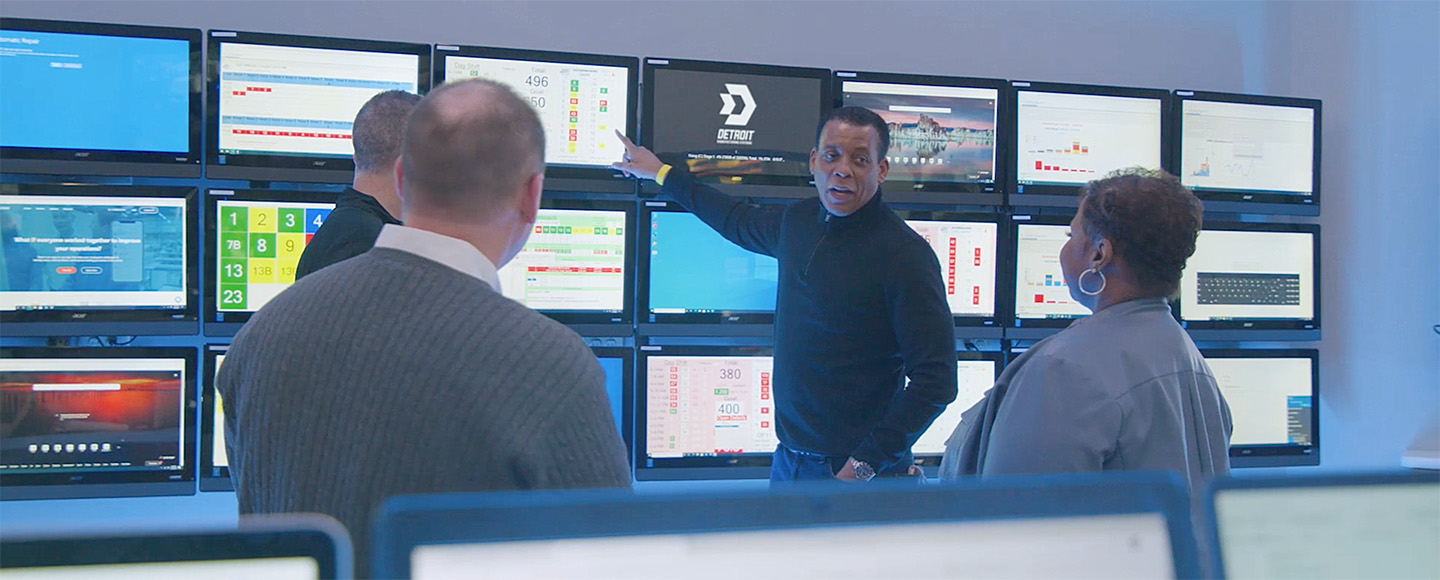 A group of people stand around a wall of tv screens while a man points at information on a screen.