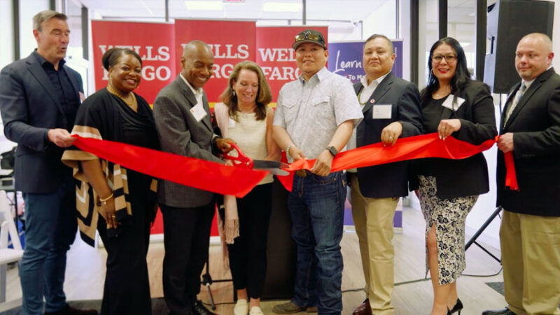 Several people stand behind a ribbon as a person near the center cuts the ribbon.