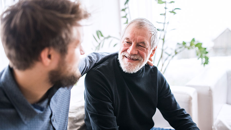 An older adult sits on a couch smiling. A younger adult sits next to him with his arm around him.