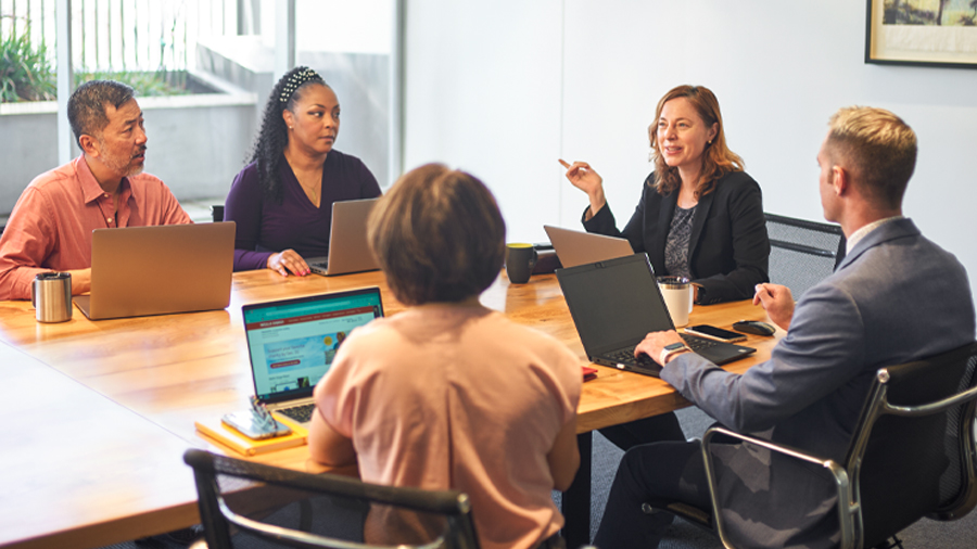 Five people sit around a conference table, each with a laptop in front of them.