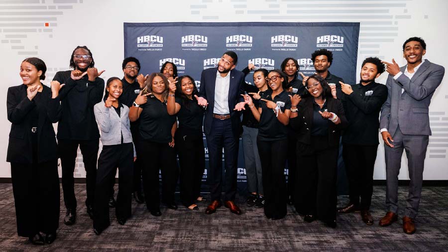 A dozen or so smiling college students point toward a professional football player in the center of the group.