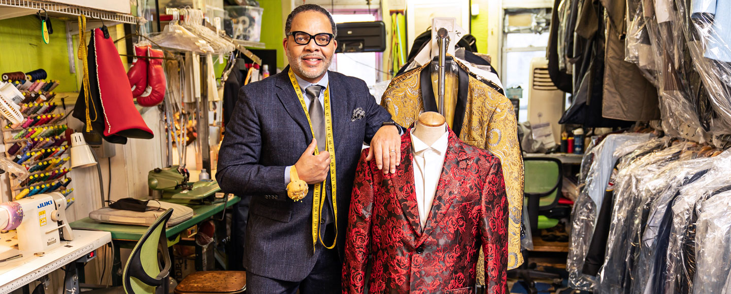 Eddie Lofton stands in his shop, JC Lofton Tailors, next to a mannequin with a suit and a rack of clothes in plastic wrap.