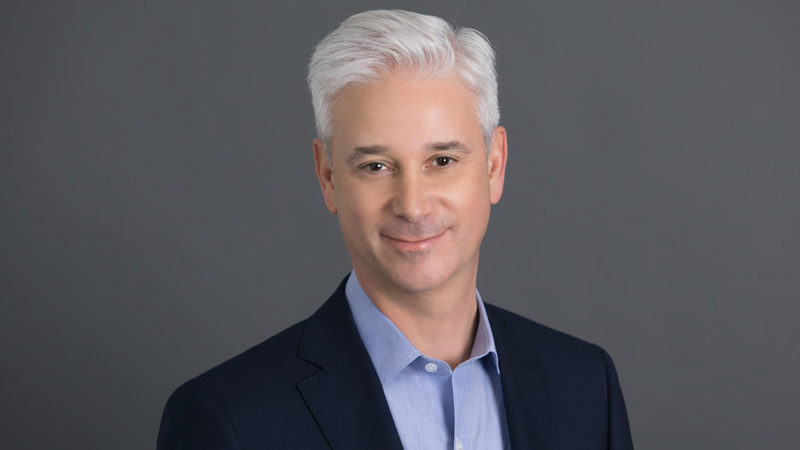 Charles Scharf, in a blue shirt and black blazer, smiling for the camera.