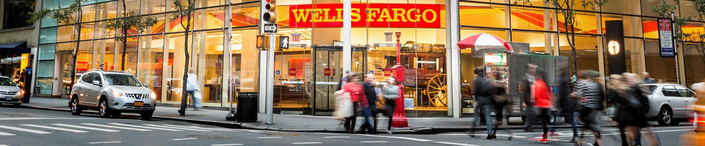 A Wells Fargo retail branch with lots of glass windows is shown in the background, with a street in front. People and cars are moving on the street.